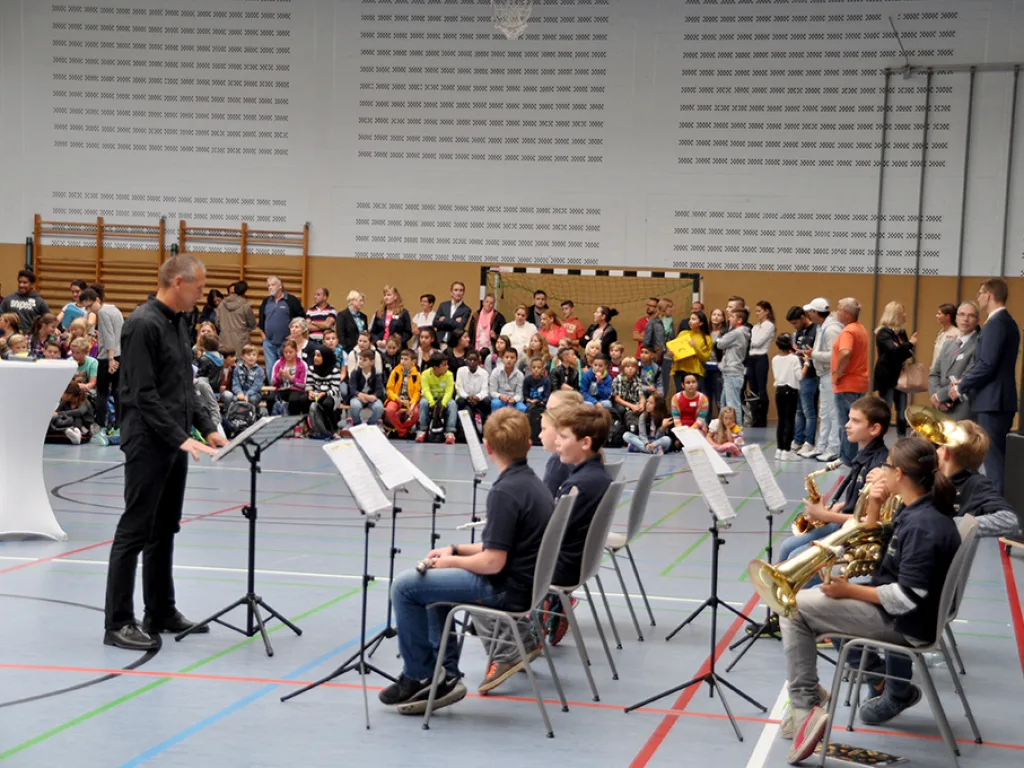 Ein Dirigent leitet eine kleine Musikgruppe in der Sporthalle.